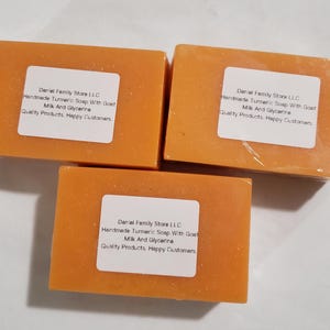 Puede incluir: Tres barras rectangulares de jabón de cúrcuma naranja, envueltas en plástico transparente con una etiqueta blanca. El texto dice: Daniel Family Store LLC, Handmade Turmeric Soap With Goat Milk And Glycerine, Quality Products, Happy Customers.