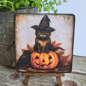May include: A square wooden sign on a small easel featuring an illustration of a black cat wearing a witch hat, sitting on an orange jack-o'-lantern surrounded by autumn leaves, with a black crow standing to the left of the pumpkin.