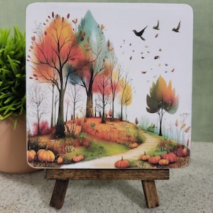 May include: A square coaster featuring a watercolor illustration of an autumn forest scene with orange, red, and yellow trees, a winding path, scattered pumpkins, and birds flying in the sky, displayed on a small wooden easel stand.