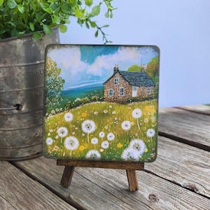 May include: A square wooden plaque featuring a painted landscape of a stone cottage nestled in a field of white dandelion seed heads under a blue sky. The artwork is displayed on a small wooden easel, showcasing detailed brushwork and colors.