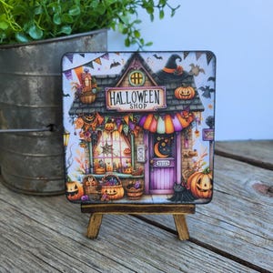May include: A square coaster featuring a colorful illustration of a Halloween shop with a purple door, orange pumpkins, hanging banners, and bats. The sign above the shop reads Halloween Shop. The coaster is displayed on a small wooden easel.