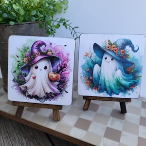 May include: Two square coasters featuring watercolor illustrations of ghosts wearing witch hats. The left ghost wears a purple hat and holds a pumpkin, while the right ghost wears a blue hat adorned with orange flowers, both set against colorful backgrounds.