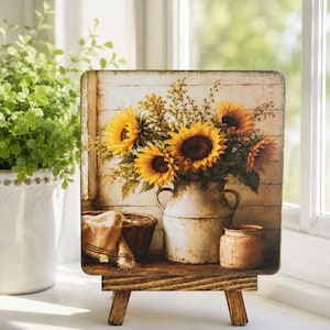 May include: A square decorative sign featuring a printed image of a bouquet of yellow sunflowers in a white metal pitcher, a woven basket with a cloth, and a small jar, all resting on a wooden stand with four short legs.