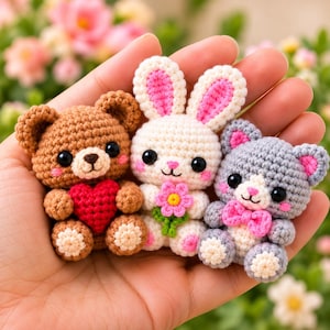 May include: Three small crocheted amigurumi figures: a brown bear holding a red heart, a white rabbit with pink-lined ears holding a pink flower, and a gray cat with a pink bow tie. Each figure features black safety eyes and stitched details.