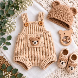 May include: A beige crochet romper with two wooden buttons and a bear face patch on the front pocket, a matching bear-eared beanie, a pair of bear-faced booties, and a wooden bear-shaped rattle, all featuring a textured, hand-stitched appearance.