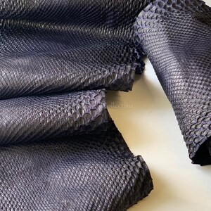 Puede incluir: Una piel de serpiente de cuero azul marino oscuro con una textura de escamas realzada y distintiva. El material est&aacute; doblado en varias capas, mostrando el patr&oacute;n repetitivo en forma de diamante en la superficie. El texto MatelsTanneryID es visible.