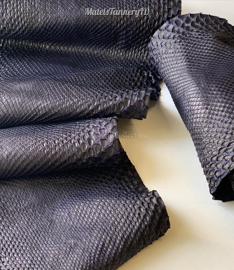 Puede incluir: Una piel de serpiente de cuero azul marino oscuro con una textura de escamas realzada y distintiva. El material est&aacute; doblado en varias capas, mostrando el patr&oacute;n repetitivo en forma de diamante en la superficie. El texto MatelsTanneryID es visible.