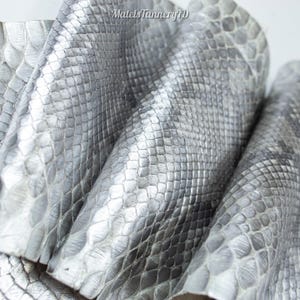 Peut inclure: Une pièce de cuir argenté métallique avec un motif texturé en peau de serpent. Le matériau présente un design à écailles répétitives avec une finition réfléchissante. Le texte MatelsTanneryID est imprimé en haut au centre.