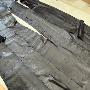 May include: Long, dark charcoal gray snakeskin hides with a distinct, repeating scale pattern. The leather pieces are laid out flat, showing a smooth, elongated texture across the surface. The text MatelsTanneryID is visible at the top.
