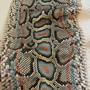 May include: A piece of snakeskin leather featuring a repeating scale pattern in shades of black, white, light blue, and orange. The material displays a textured surface with a distinct organic design, showcasing a vibrant colour palette throughout.