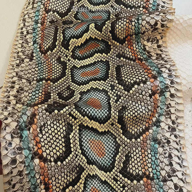 May include: A piece of snakeskin leather featuring a repeating scale pattern in shades of black, white, light blue, and orange. The material displays a textured surface with a distinct organic design, showcasing a vibrant colour palette throughout.