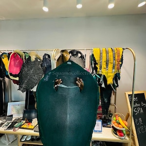 May include: A large, dark green stingray leather hide with a textured, pebbled surface and a central white calcified spot. The hide is held up, displaying its distinct oval shape and natural grain pattern, surrounded by various other leather pieces.