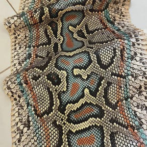 May include: A flat piece of snakeskin leather featuring a repeating scale pattern in shades of cream, black, turquoise, and burnt orange. The surface displays a natural, textured organic design with a central vertical motif and the text MatelsTannerID.