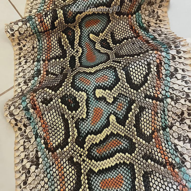 May include: A flat piece of snakeskin leather featuring a repeating scale pattern in shades of cream, black, turquoise, and burnt orange. The surface displays a natural, textured organic design with a central vertical motif and the text MatelsTannerID.