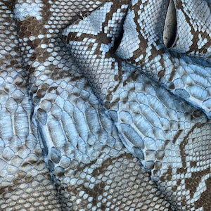 May include: A sheet of light blue and dark brown snakeskin leather featuring a repeating scale pattern and the text MatelsTannery10 printed in the upper centre. The material is folded, showing the texture and depth of the reptile skin pattern.