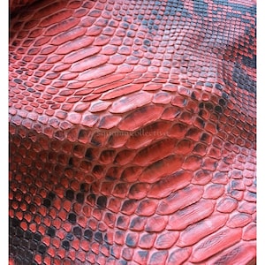 Op de afbeelding: Een getextureerd vel slangenleer in rood en zwart met een herhalend schubbenpatroon. Het oppervlak toont een levendige rode basiskleur met donkere zwarte schaduwen over de schubben. De tekst MatelsTanneryID en SquamaCollective is zichtbaar.