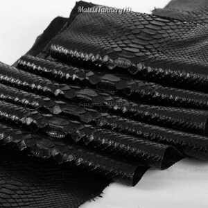 May include: A stack of black snakeskin leather sheets with a distinct, raised scale texture. The surface features a glossy finish and a repeating pattern of overlapping scales. The text MatelsTanneryID and SquamaCollection are visible on the leather.
