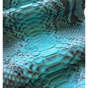 Op de afbeelding: Een stuk gestructureerd slangenleer met een levendig turquoise en zwart kleurverloop. Het oppervlak vertoont een herhalend schubbenpatroon met natuurlijke variaties in kleur en diepte. De tekst MatelsTanneryID is zichtbaar.