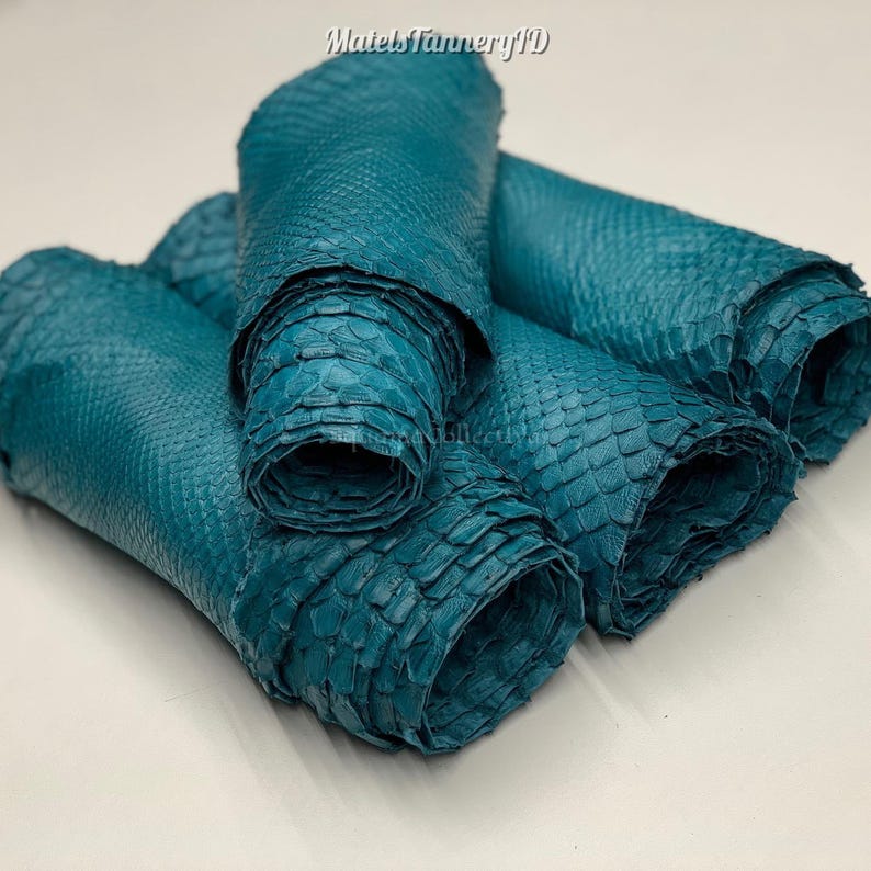 Puede incluir: Varios rollos de piel de serpiente de color verde azulado con una textura de escamas en relieve. El material es flexible y muestra un tono azul verdoso profundo y uniforme en toda la superficie. El texto MatelsTanneryID es visible arriba.