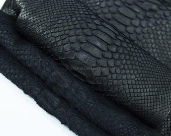 Puede incluir: Una pieza de cuero negro doblada con un patrón texturizado de piel de serpiente. La superficie muestra un diseño de escamas repetitivas con un acabado mate. El texto MatelsTanneryID y SquamaCollectiva es visible en el material.