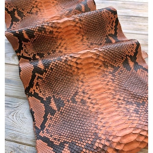 Peut inclure: Une pièce de cuir de serpent orange et noir présentant un motif d'écailles distinct. Le matériau est plié en couches, affichant une surface texturée avec un dégradé de marques anthracite foncé sur une base orange vif. Le texte indique MatelsTanneryID.