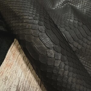 May include: A dark charcoal black snakeskin leather hide featuring a distinct, overlapping scale pattern across the surface. The material displays a matte finish with visible texture and natural variations in the scale size and shape throughout.