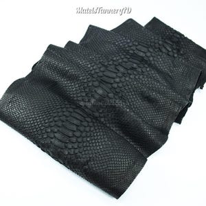 Peut inclure: Une pile de feuilles de cuir de serpent noir présentant un motif d'écailles reptiliennes distinct. La couche supérieure affiche une rangée centrale proéminente d'écailles allongées flanquées d'écailles texturées. Le texte MatelsTanneryID est visible.