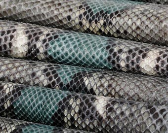 Puede incluir: Una lámina enrollada de piel de serpiente con un patrón repetitivo de escamas grises, negras y verde azulado. La superficie muestra una estructura orgánica texturizada con la marca de agua MatelsTanneryID visible.