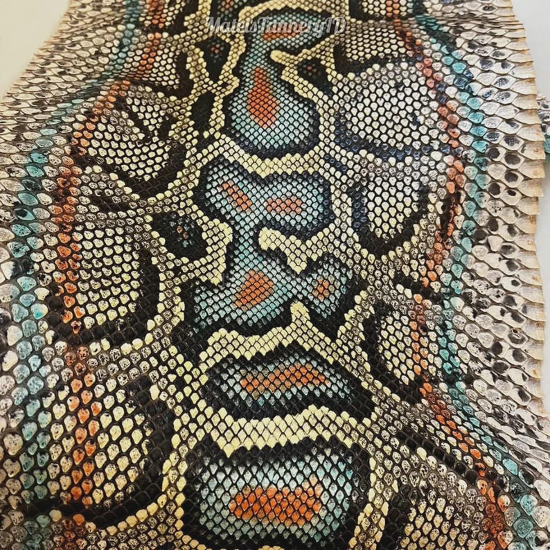May include: A piece of snakeskin leather featuring a repeating scale pattern in shades of black, white, cream, turquoise, and orange. The text "MatelsTanneryID" is printed in a small, light-coloured font near the top centre of the material.