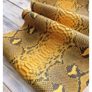 May include: A piece of yellow and black snakeskin leather featuring a distinct scale pattern and the text MatelsTanneryID printed at the top. The material is folded over itself, showcasing the textured surface and the natural organic design.