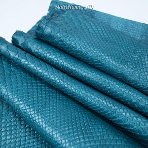 Puede incluir: Una pieza doblada de piel de serpiente de color verde azulado que presenta una textura de escamas en relieve en toda la superficie. El texto MatelsTanneryID es visible en la parte superior de la imagen, impreso en una fuente simple y oscura.