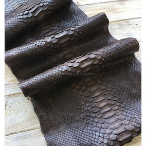 Op de afbeelding: Een donkerbruine slangenleren huid met een opvallend schubbenpatroon met grotere, langwerpige schubben in het midden en kleinere, fijnere schubben langs de randen. Het materiaal is in lagen gevouwen en heeft een matte afwerking.