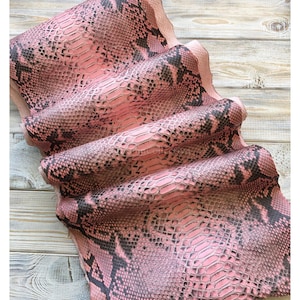May include: A folded piece of leather featuring a pink and black snakeskin pattern. The surface has a textured, scale-like appearance. The text "MatelsTanneryID" is visible at the top of the image, printed in a simple font.