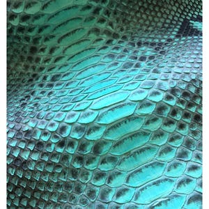 Op de afbeelding: Een vel echt slangenleer met een levendig turquoise en zwart schubbenpatroon. Het oppervlak vertoont een natuurlijke, herhalende textuur met de tekst MatelsTanneryID in een klein, duidelijk lettertype bovenaan in het midden gedrukt.