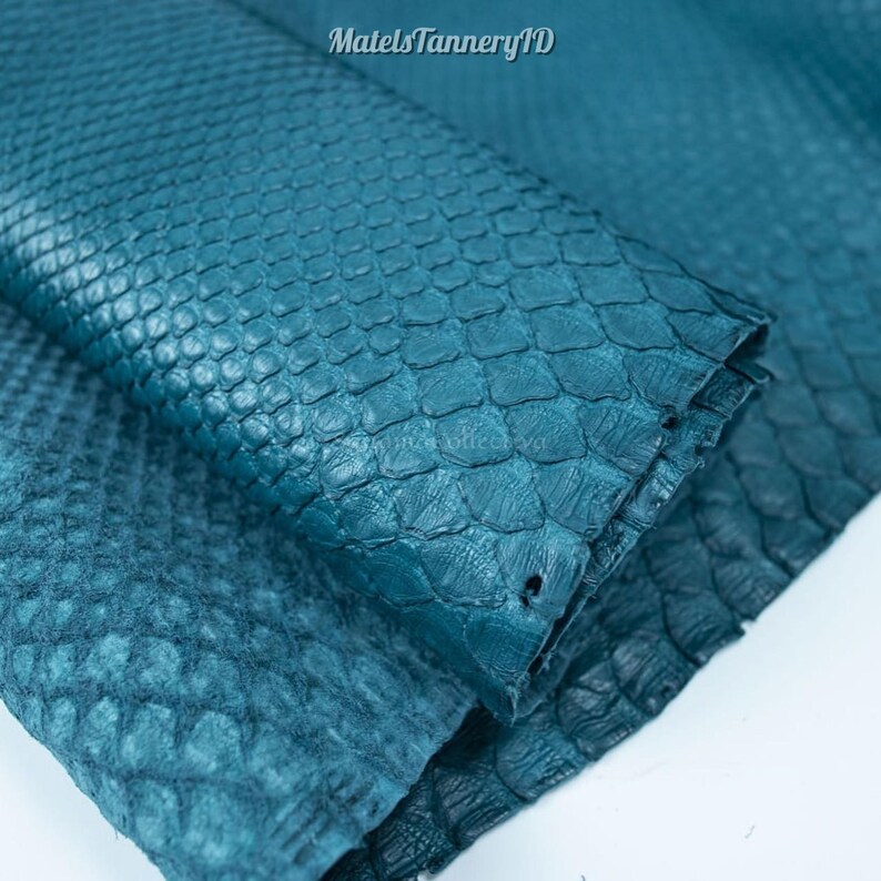 Puede incluir: Una pieza de cuero de serpiente texturizado de color verde azulado con un patr&oacute;n de escamas repetitivo y distintivo en toda la superficie. El material est&aacute; doblado sobre s&iacute; mismo, mostrando la textura org&aacute;nica superpuesta. El texto MatelsTanneryID es visible.