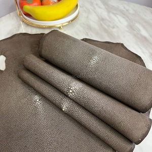 Könnte beinhalten: Drei Rollen aus braunem Rochenleder mit einer ausgeprägten, körnigen Textur und einem zentralen weißen, kreisförmigen Muster. Das Leder liegt auf einem flachen, unregelmäßigen Stück derselben braunen Rochenhaut mit sichtbarer Maserung.