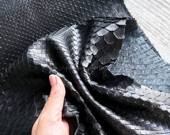 Op de afbeelding: Een stuk zwart slangenleer met een duidelijk, overlappend schubbenpatroon. Het materiaal heeft een glanzende afwerking en is gevouwen om de flexibiliteit te tonen. De tekst MatelsTanneryID is zichtbaar aan de bovenkant van het leeroppervlak.