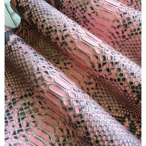Op de afbeelding: Een vel roze en zwart slangenleer met een gestructureerd, herhalend schubbenpatroon. Het materiaal is in lagen gevouwen, wat de gedetailleerde oppervlaktestructuur en de tekst MatelsTanneryID op het bovenste gedeelte laat zien.