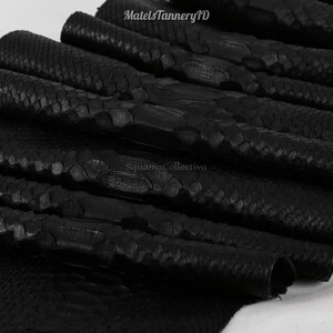 Peut inclure: Une pile de feuilles de cuir noir texturé avec un motif d'écailles distinct. Le texte MatelsTanneryID et SquamaCollectiva est visible sur la surface. Le matériau présente une finition mate et une texture d'écailles organique répétitive.