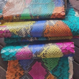 May include: A stack of multi-colored snakeskin leather sheets featuring a repeating diamond scale pattern in vibrant shades of pink, blue, yellow, orange, and green. The text MatelsTanneryID is visible at the top of the top leather sheet.