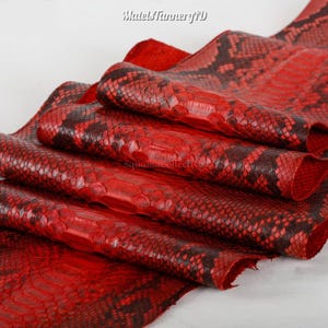 May include: A piece of red and black snakeskin leather featuring a distinct scale pattern. The material is folded in layers, showcasing the textured surface and the vibrant crimson and dark charcoal coloration. The text MatelsTanneryID is visible.