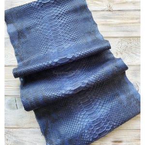 May include: A folded piece of blue snakeskin leather featuring a distinct scale pattern and a gradient of dark blue to black tones. The text MatelTanneryID and SquamaCollection are visible on the surface of the textured material.