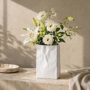 Puede incluir: Un jarrón de cerámica blanca con una superficie texturizada que recuerda al papel arrugado contiene un ramo de lirios blancos y flores de eustoma. El jarrón tiene una forma rectangular con pliegues y arrugas irregulares en su exterior.