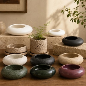 Puede incluir: Una colección de varios maceteros y cuencos de cerámica en diversas formas, texturas y colores, incluyendo blanco mate, beige moteado, gris oscuro, verde bosque y ciruela intenso, con un macetero cilíndrico de piedra de textura rugosa.