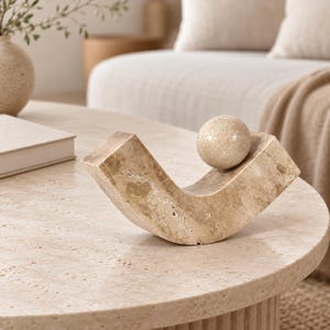 May include: A beige travertine stone sculpture featuring a curved, angular base with a porous, textured surface and a smooth, spherical ball resting on one side of the structure. The object displays natural tan and cream colour variations throughout.