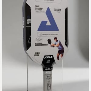 Puede incluir: Una pala de pickleball Joola Ben Johns Hyperion C2 negra con una funda de cartón blanca. La funda presenta marca azul, texto que incluye 16mm Charged Carbon y la imagen de un jugador. El mango tiene una envoltura blanca con texto.