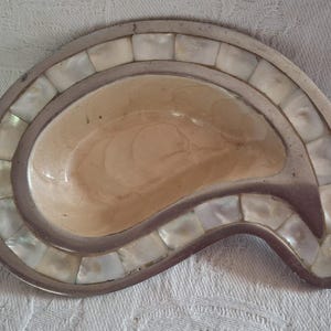 May include: A paisley-shaped decorative dish featuring a smooth, cream-coloured central basin surrounded by a border of inlaid, iridescent square mother-of-pearl tiles set within a dark, metallic-toned frame with a curved, asymmetrical silhouette.