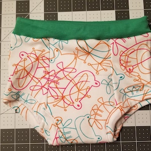 May include: White baby underwear with a green waistband. The underwear is patterned with orange, pink, and teal abstract shapes. The fabric is soft and appears to be cotton.