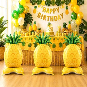 May include: Three large pineapple-shaped foil balloons stand in the foreground. In the background, a table is decorated with a yellow fringe skirt, a pineapple-themed tablecloth, a birthday cake, and a banner that reads Happy Birthday.