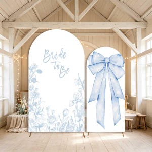 Puede incluir: Dos paneles de fondo arqueados para eventos con fondo blanco y dibujos lineales florales en azul claro. El panel más grande muestra el texto "Bride to be" en letra cursiva, mientras que el más pequeño presenta un gran lazo azul claro.